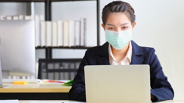 A person wearing a face mask is seated at a desk, focusing on their laptop with a shelf of books in the background.