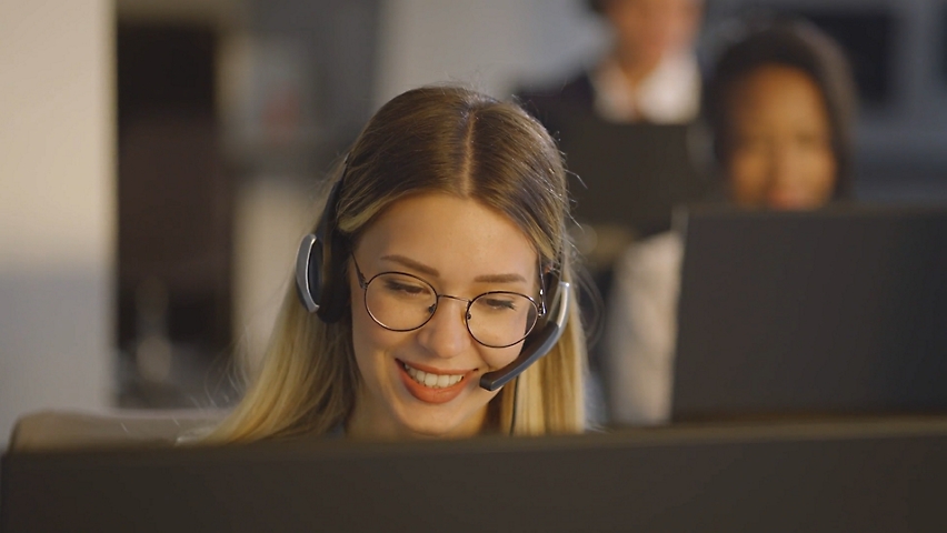 Ženska s slušalkami sedi v pisarni pred namiznim računalnikom in govori