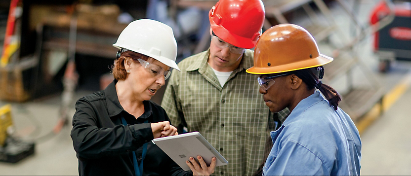 Three people wearing hard hats and safety glasses stand in a workplace, examining a tablet