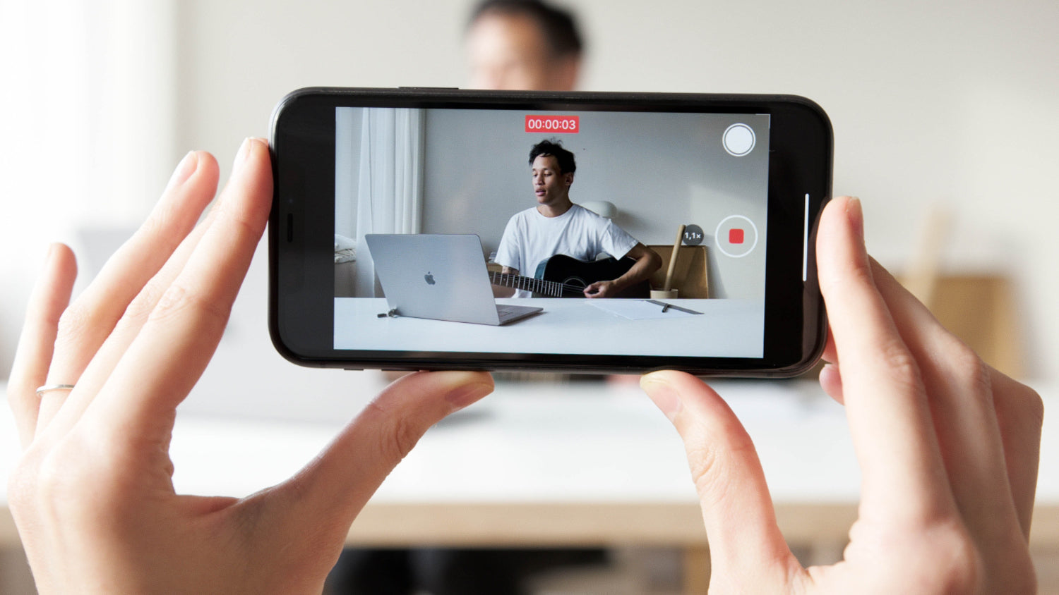 Two hands in the foreground hold a phone that is recording a musician