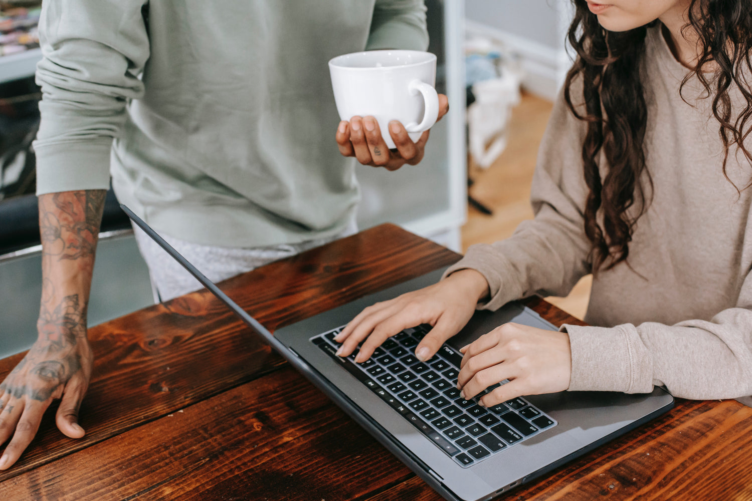 Two people work together on a laptop. One holds a coffee