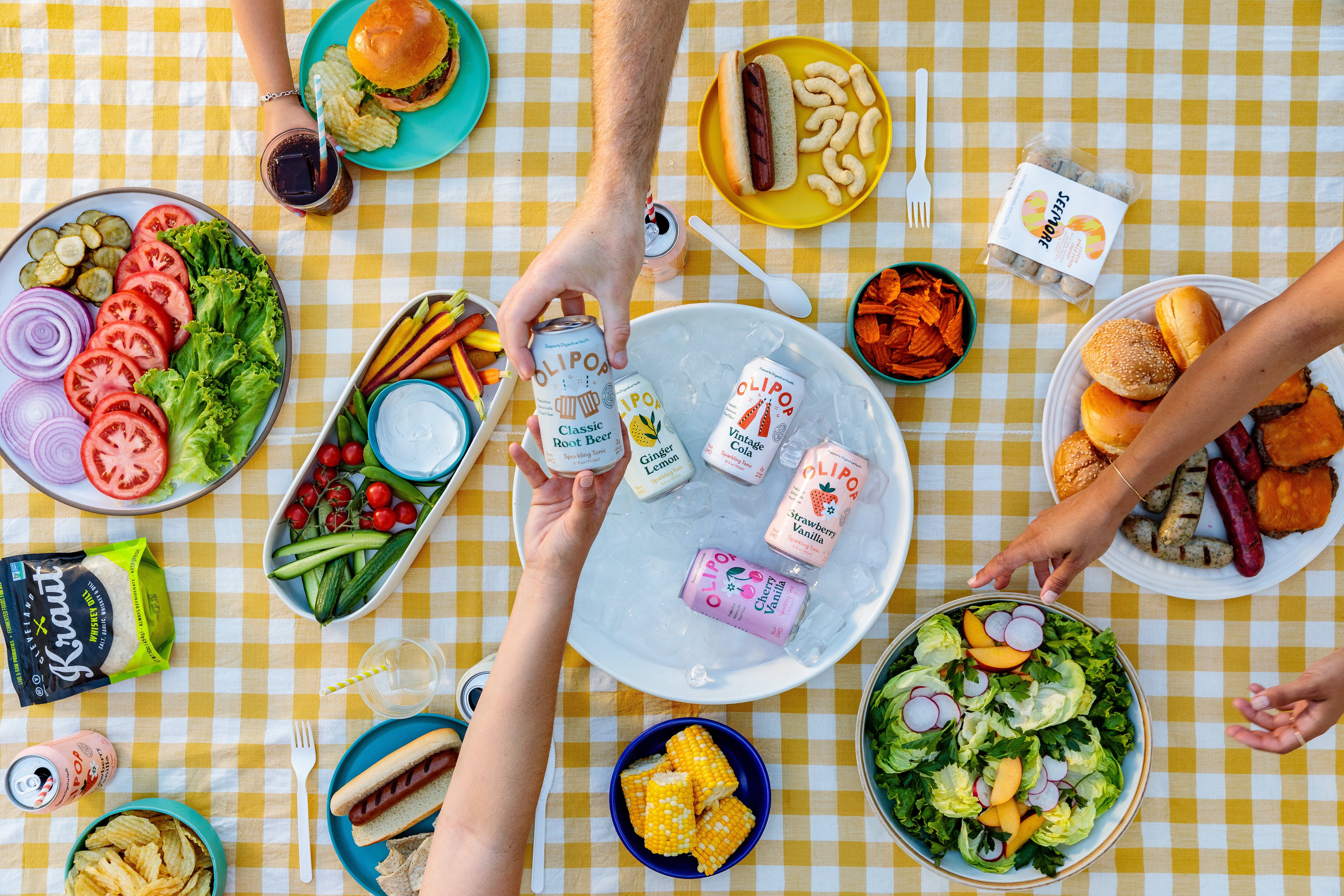 An aerial shot of a flatlay picnic set up, with hands passing a can of Olipop