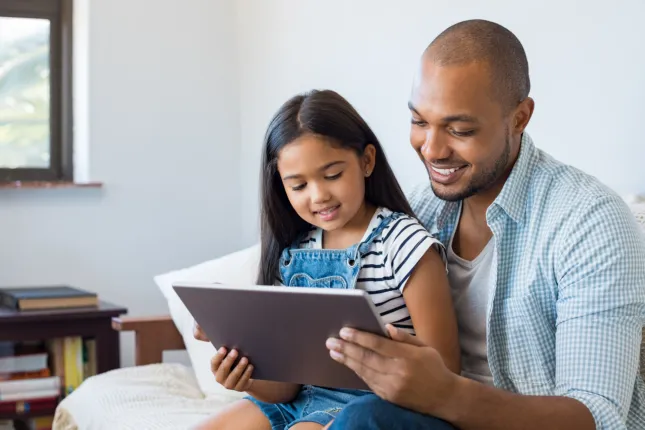 Un monsieur souriant et une jeune enfant regardent une tablette. 
A smiling man and a young lady looking at a tablet.