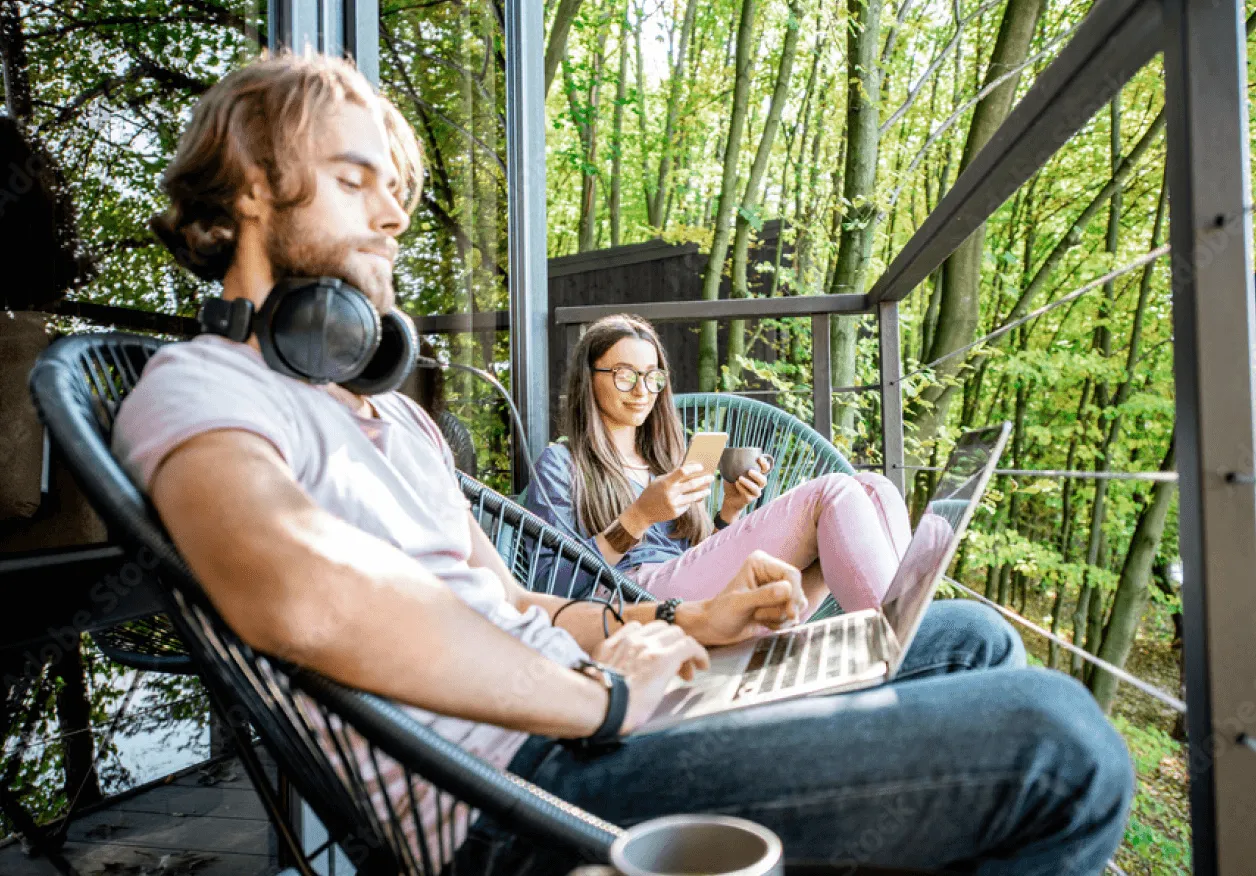 Two young adults sit outside their home, surrounded by nature, both using wireless devices.