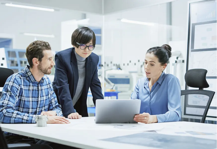 One standing and two sitting colleagues looking at a laptop computer