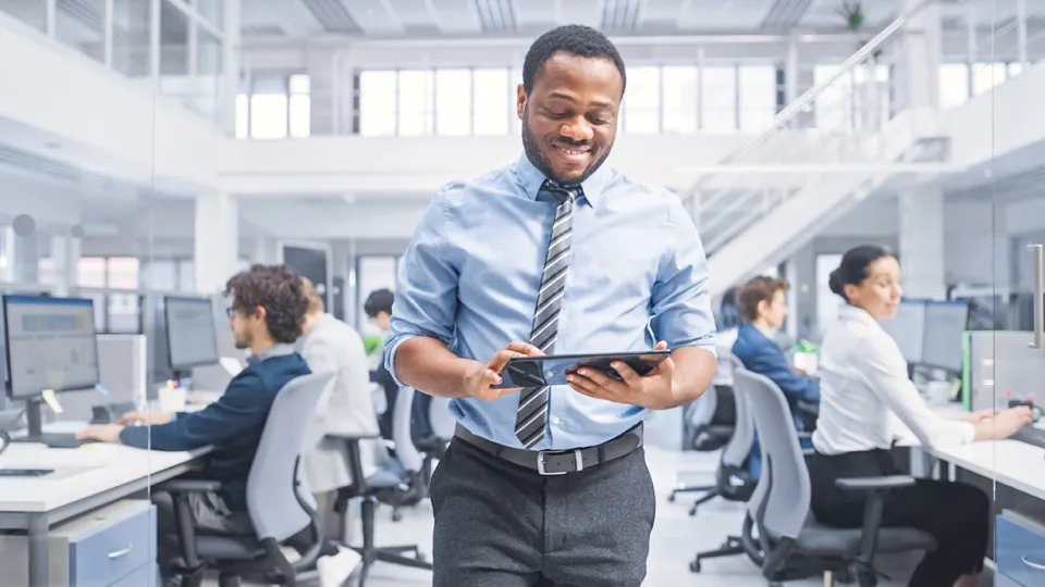 Smiling man walking through an open plan office and working on a digital tablet