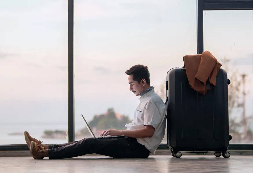 a man sitting on the floor with a laptop with his back resting against some travel bags at the airport