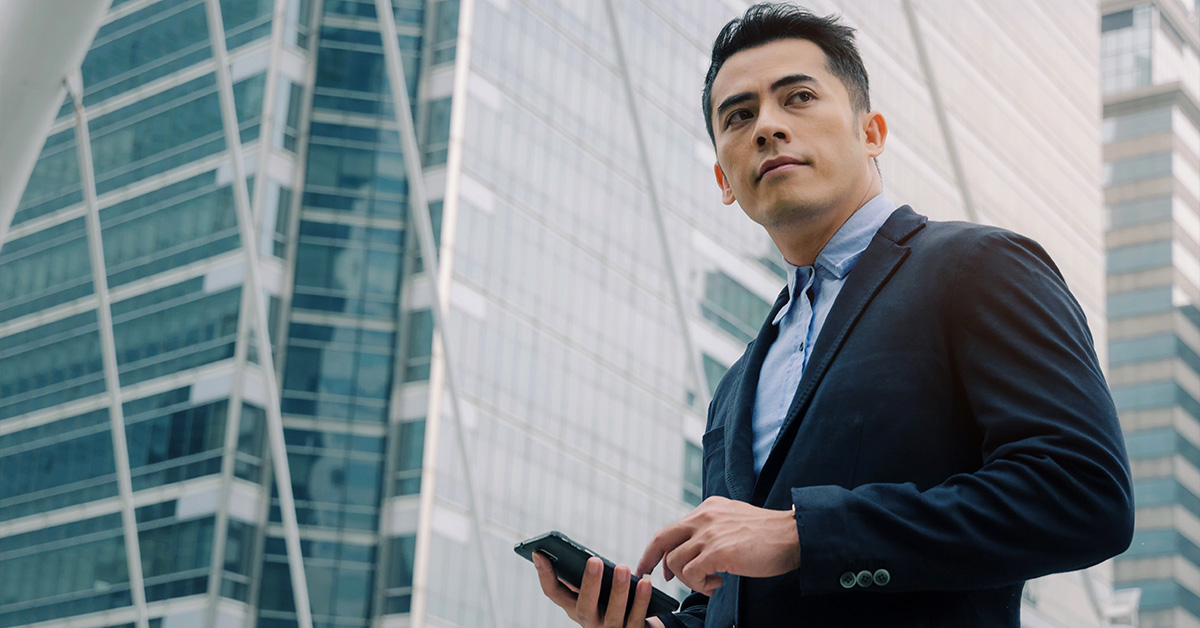 Man in suite looking down at his phone with large buildings in the background.