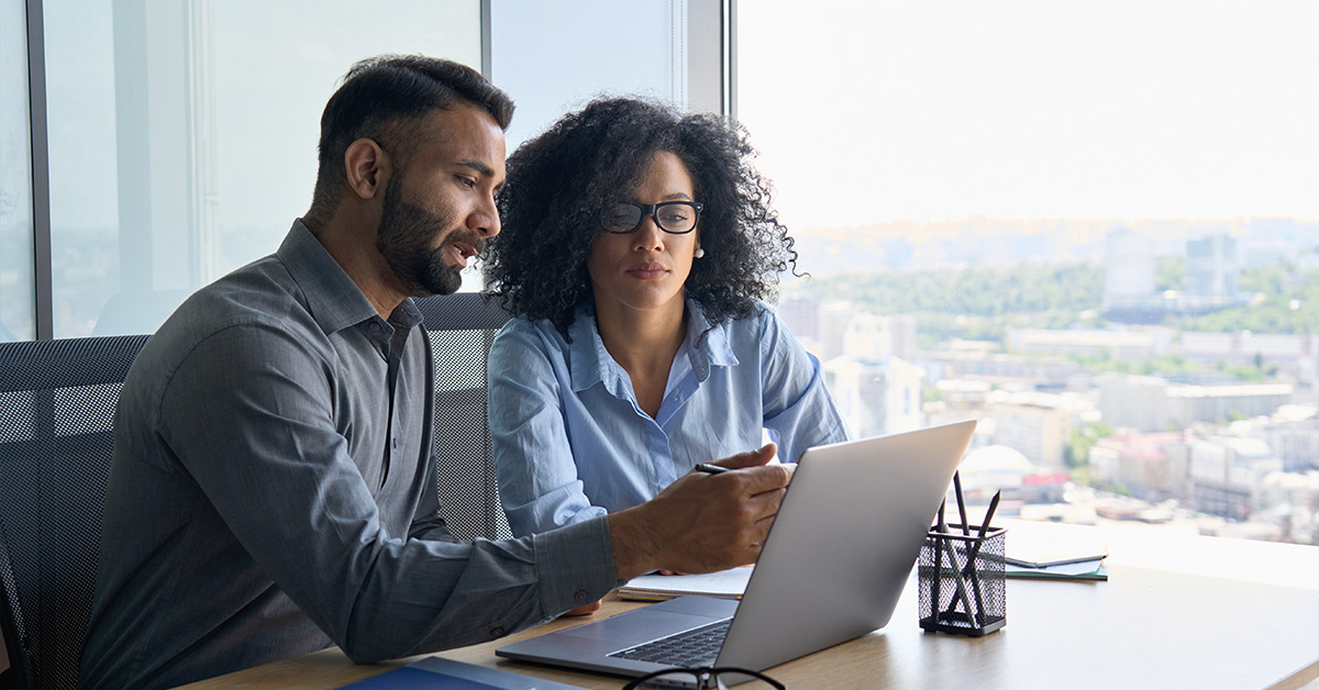 Man and woman in modern office looking and pointing at laptop in front of them.