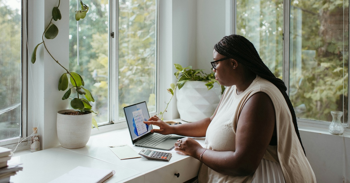 A woman works from home at her desk by the window, tapping the touch screen on her laptop.