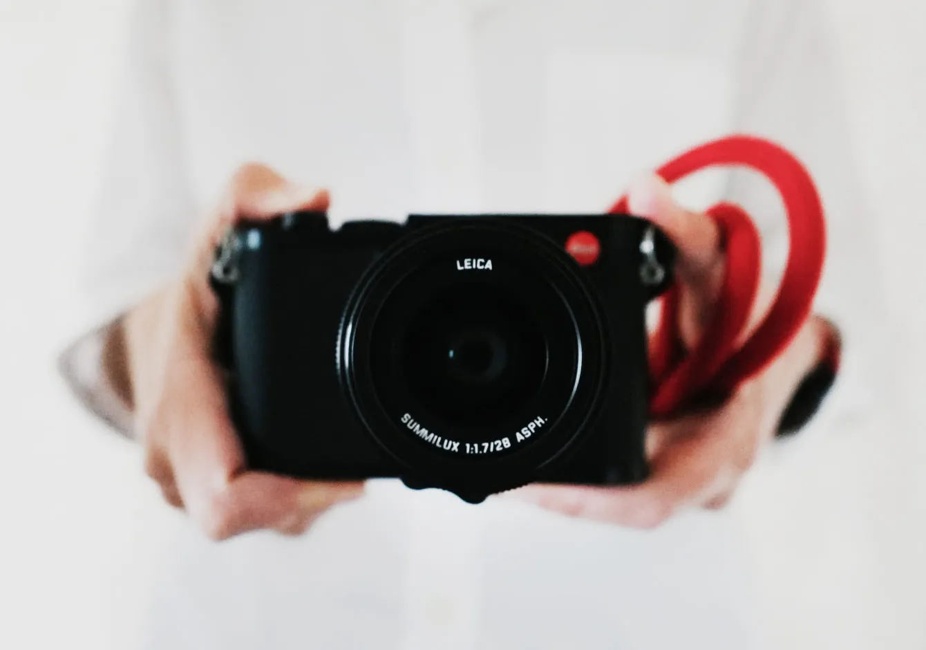 Closeup of photographer's hands holding a Leica camera with a red strap on a blurred white background