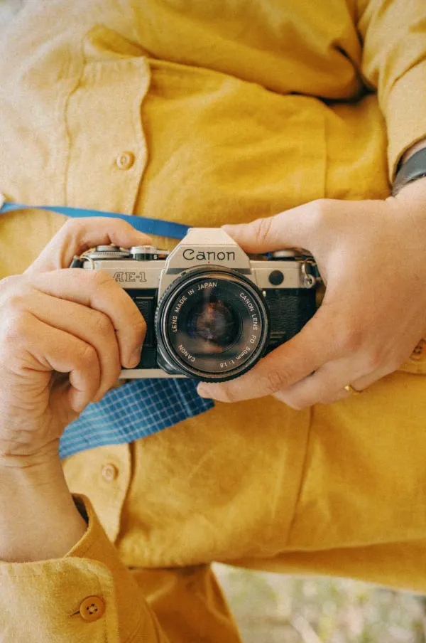 Photographer's hands and chest wearing a yellow shirt taking a selfie on a Canon AE-1 camera with a blue strap