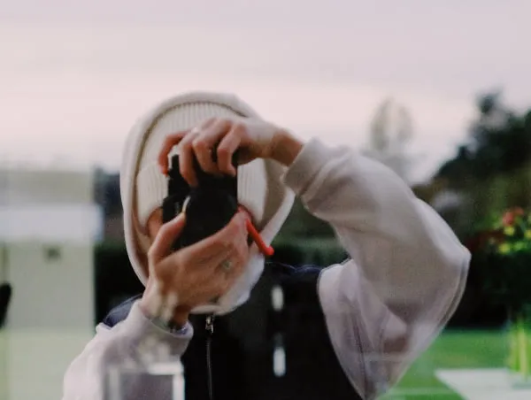 Photographer taking selfie in a window reflect behind camera lens wearing a white hood and white beanie