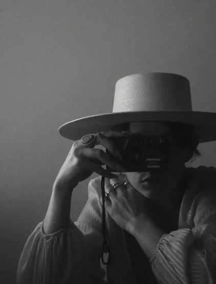 Black and white self portrait of photographer behind a camera lens wearing a wide brim hat and rings