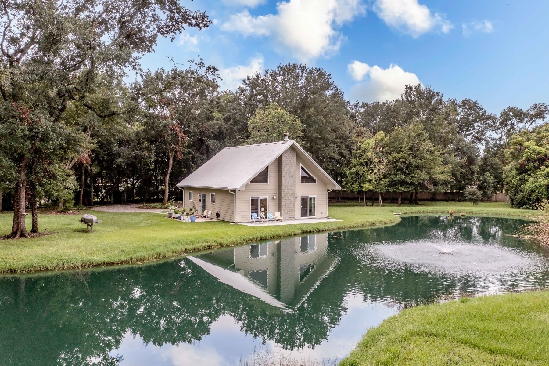 A house with a sloped roof sits beside a tranquil pond surrounded by lush trees and greenery.