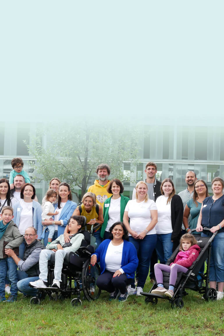 A group photo of adults and children, some of whom are in wheelchairs, gathered outdoors with smiles set against a soft blue background.