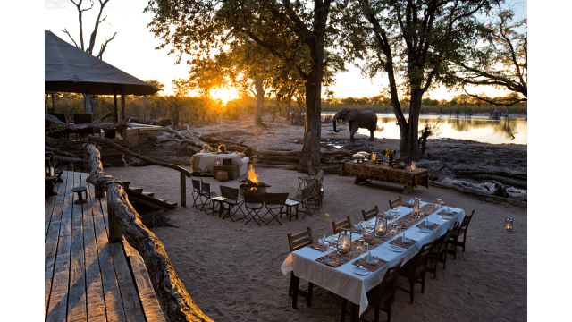 Tourists sit watching an elephant drink at a waterhole in front of a safari lodge.