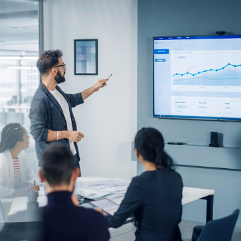 Young man giving a presentation to coworkers that are sitting at a desk and he is pointing to a monitor on the wall