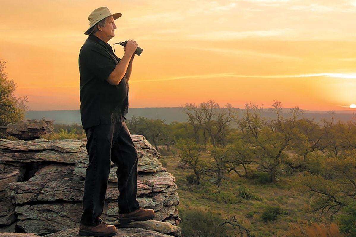 A man in a hat and boots holds binoculars while standing on a rocky point looking over brushland and a sunset.