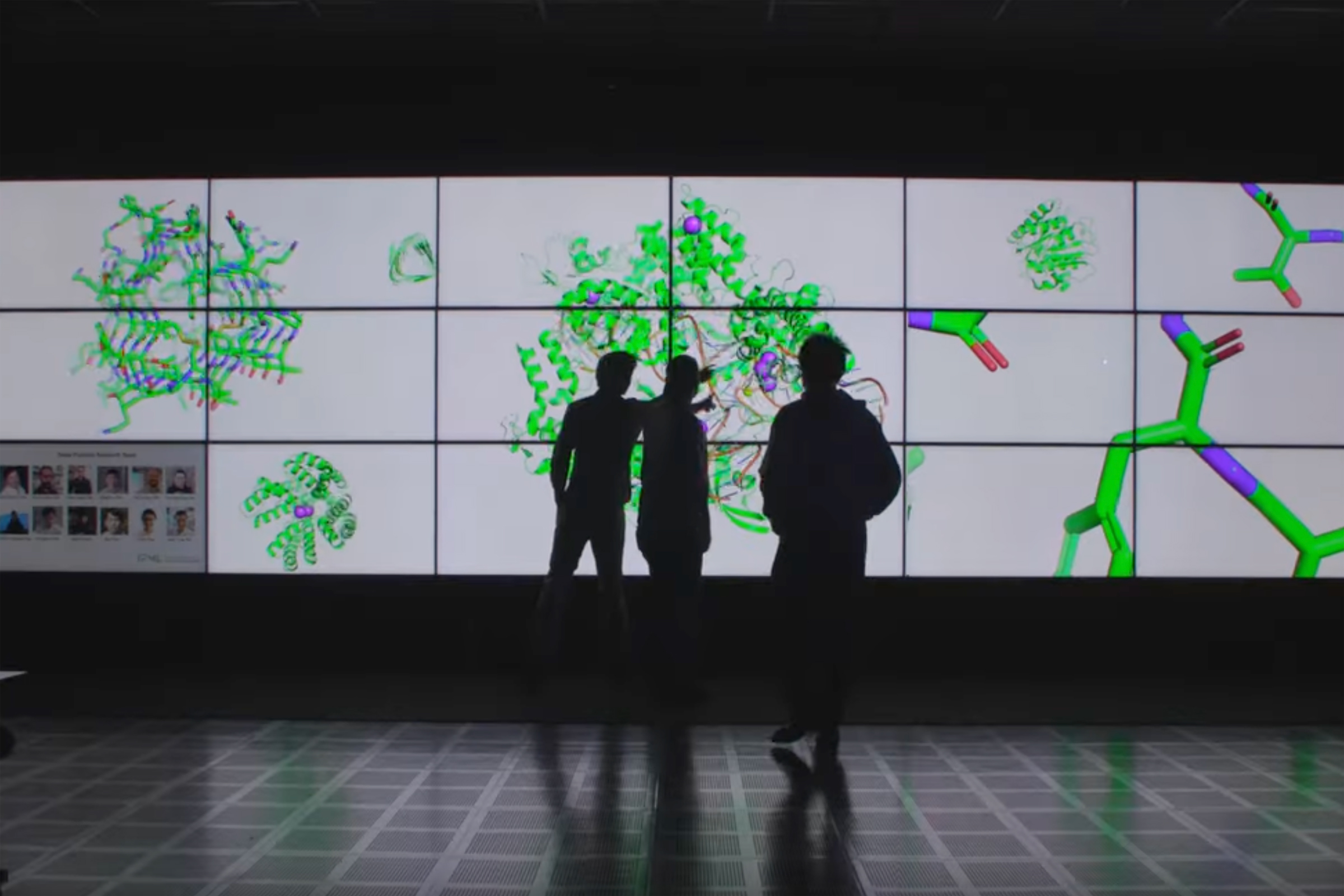 Three people stand silhouetted  in front of a wall-sized video display that shows several large colorful illustrations of molecules