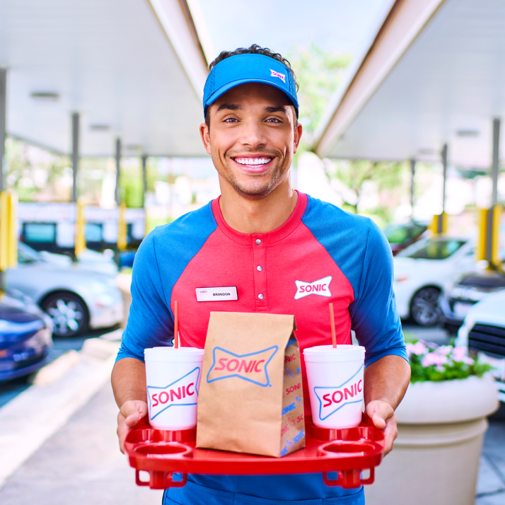 Smiling Sonic employee holding a tray with drinks and a bag.