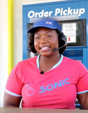 Sonic employee with headset and a red shirt, smiling at Order Pickup station