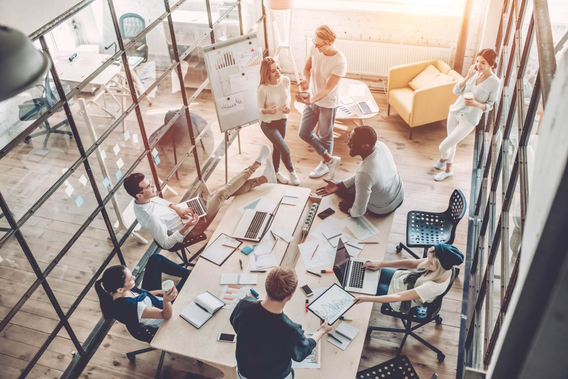 A diverse group of people working together in a modern office with large windows. They are gathered around a table with laptops, notebooks, and charts. Some are seated while others stand, interacting and discussing ideas. A whiteboard displays graphs and notes.