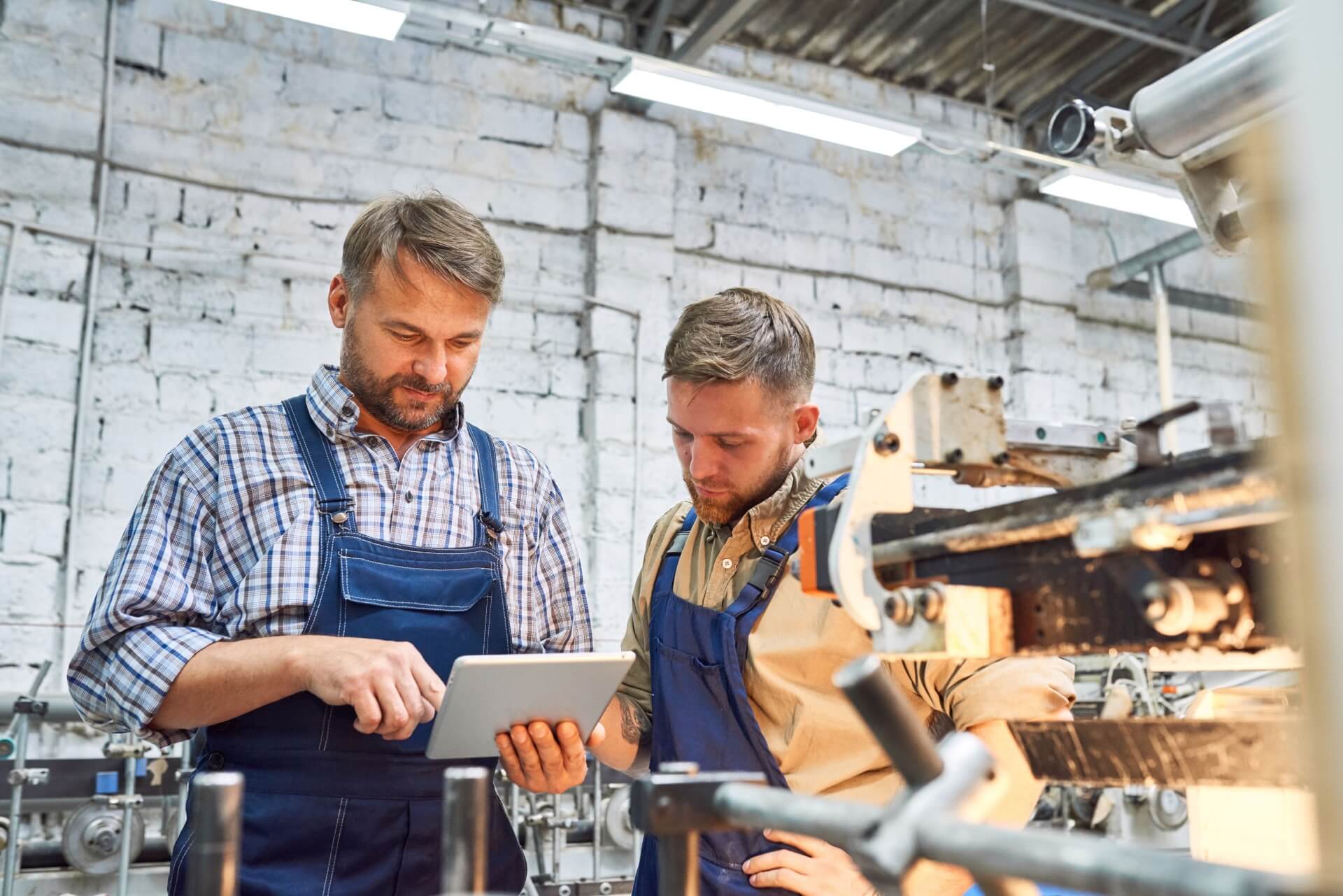 Two factory workers in blue overalls and plaid shirts study information on a tablet, showcasing the seamless integration of technology in the B2B manufacturing sector, with machinery humming efficiently in the background.