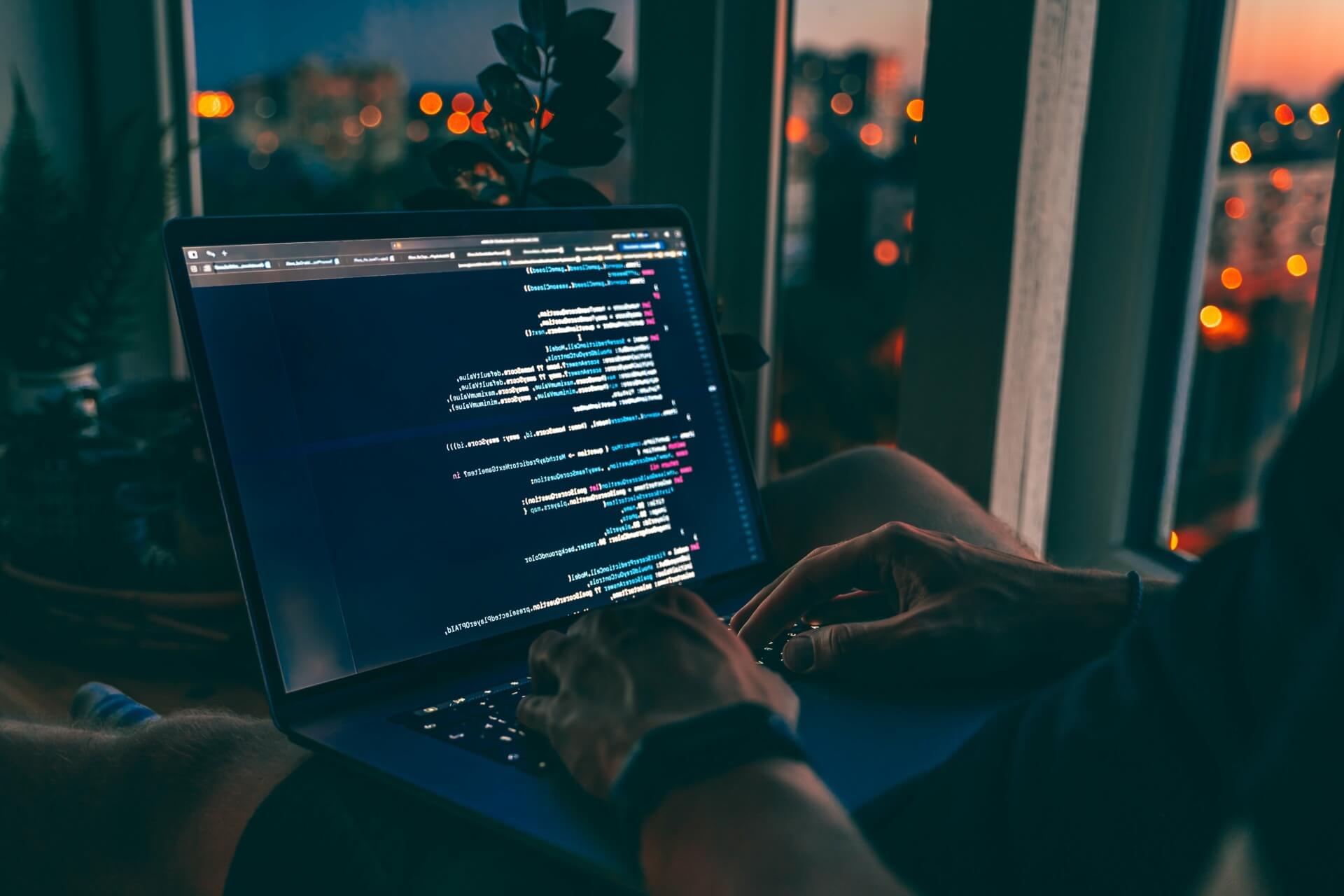 Person typing code on a laptop with a nighttime cityscape visible through a window in the background. The screen displays colorful programming syntax, reflecting an ecosystem-led innovation approach.