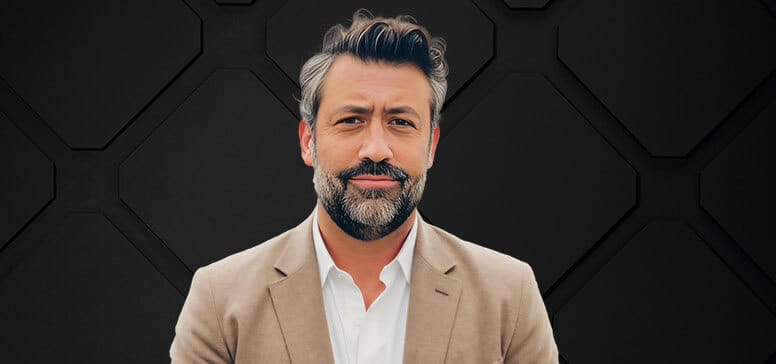 A man with grey hair and a beard, wearing a light brown blazer and white shirt, stands in front of a textured dark background.