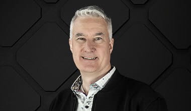 A man with short gray hair and a slight smile, exuding an air of leadership, is wearing a suit and standing in front of a dark geometric-patterned background.