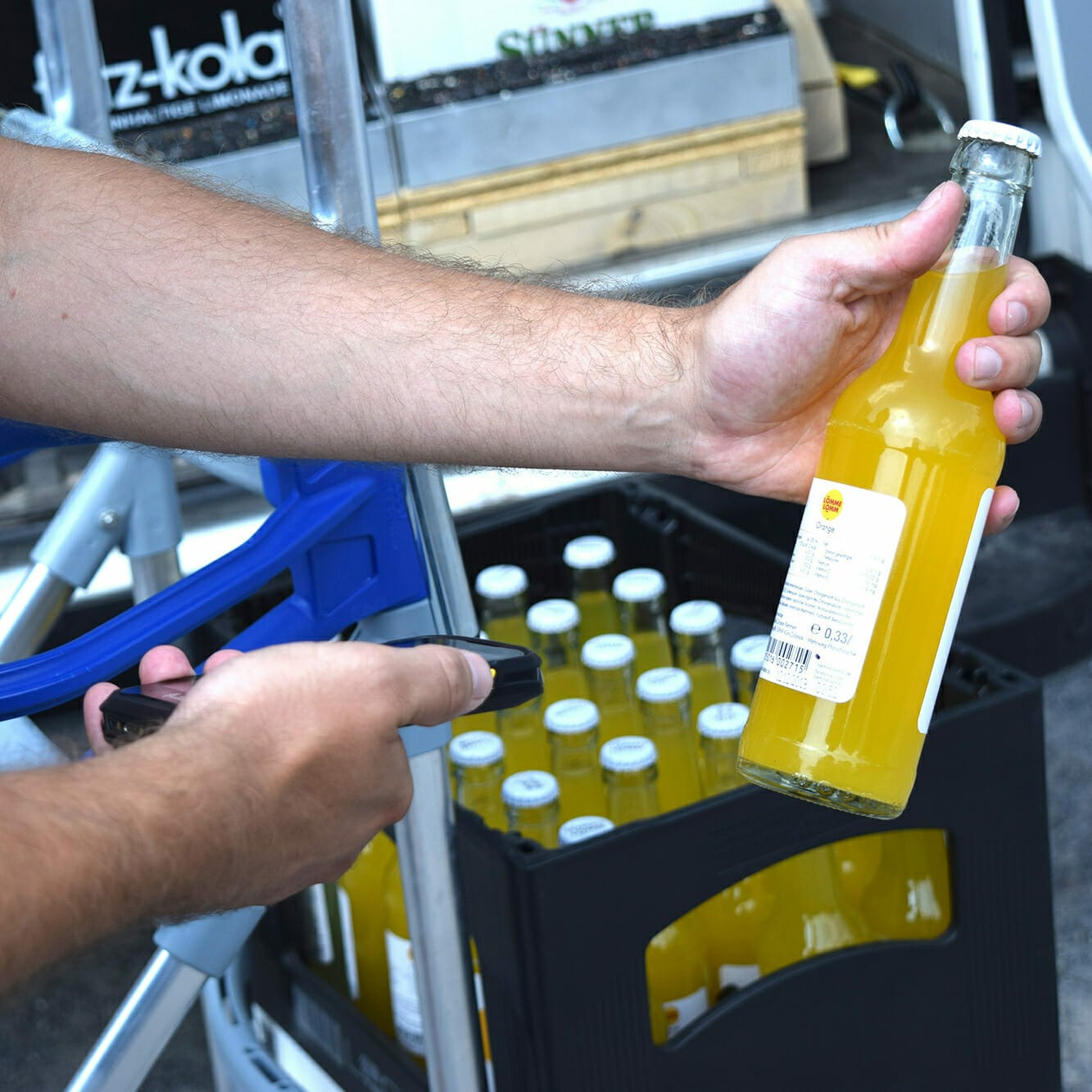 A person uses a handheld scanner to scan a bottle of yellow liquid from a crate filled with similar bottles, meticulously inspecting each as if preparing for MVP cases.