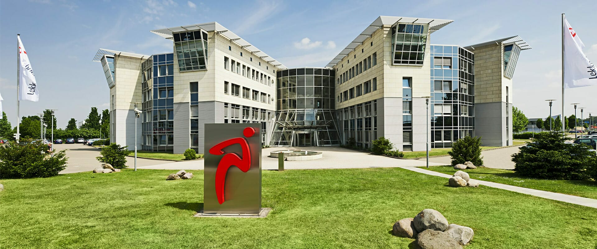 Modern office building with glass and steel architecture, fronted by a green lawn and a metal sculpture featuring the red Lekkerland logo. Flags with the same logo are visible. The sky is clear and sunny, creating an inviting environment for this wholesale giant’s headquarters.