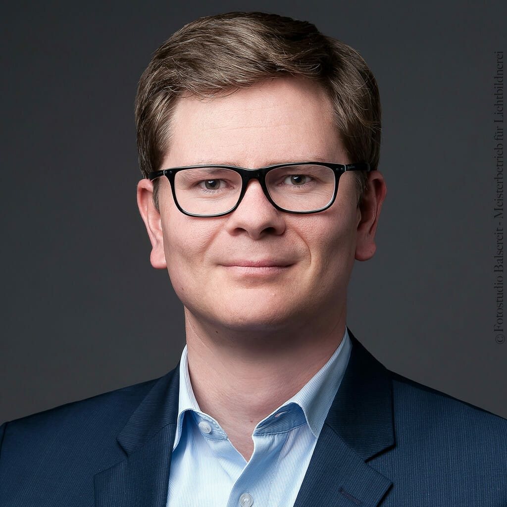 A man with short hair, wearing glasses and dressed in a blue suit with a light blue shirt, poses for a headshot against a dark grey background. This image could be perfect for Lekkerland's online wholesale platform.