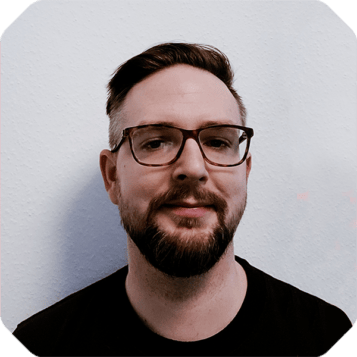 A man with short hair, a trimmed beard, and glasses wearing a black t-shirt stands against a light-colored wall, looking like he's ready to step into the central marketplace.