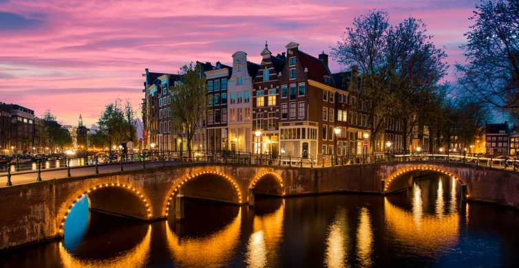 A picturesque view of Amsterdam at twilight. Historic buildings line the banks of a canal, their windows warmly lit. Arched bridges cross the water, adorned with lights that create beautiful reflections. The sky is painted in vibrant shades of pink and purple.