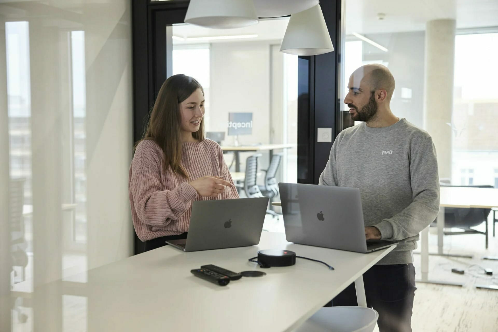 Two colleagues standing at a high table, each using a laptop, engage in conversation in a modern office, possibly discussing updates for the company's career page.