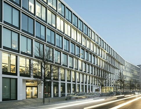 Modern multi-story office building with large windows and a minimalistic design, located along a street with light trails from passing vehicles and parked cars in the foreground, reflecting the dynamic environment suited for Spryker careers.