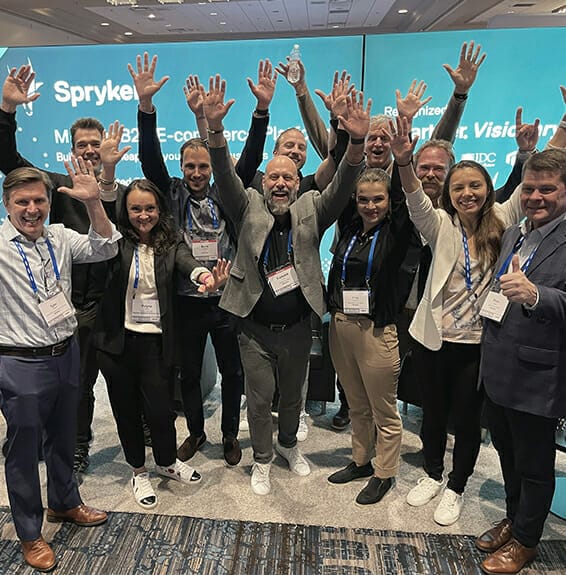 A group of people, smiling with raised hands and wearing conference badges, stands together in front of a digital display screen. They're eager to learn about Spryker's cutting-edge solutions.