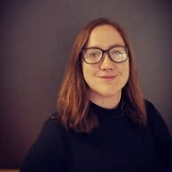 A person with shoulder-length brown hair and glasses, wearing a dark turtleneck, stands against a plain dark background, smiling slightly at the camera—a confident presence perfect for Spryker careers.