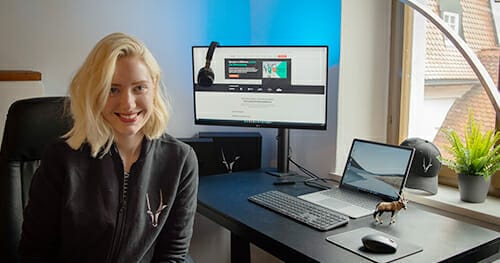 A woman with shoulder-length blonde hair sits at a desk with a computer monitor, laptop, and accessories. The monitor displays a Spryker Careers website. A window and a potted plant are visible in the background.