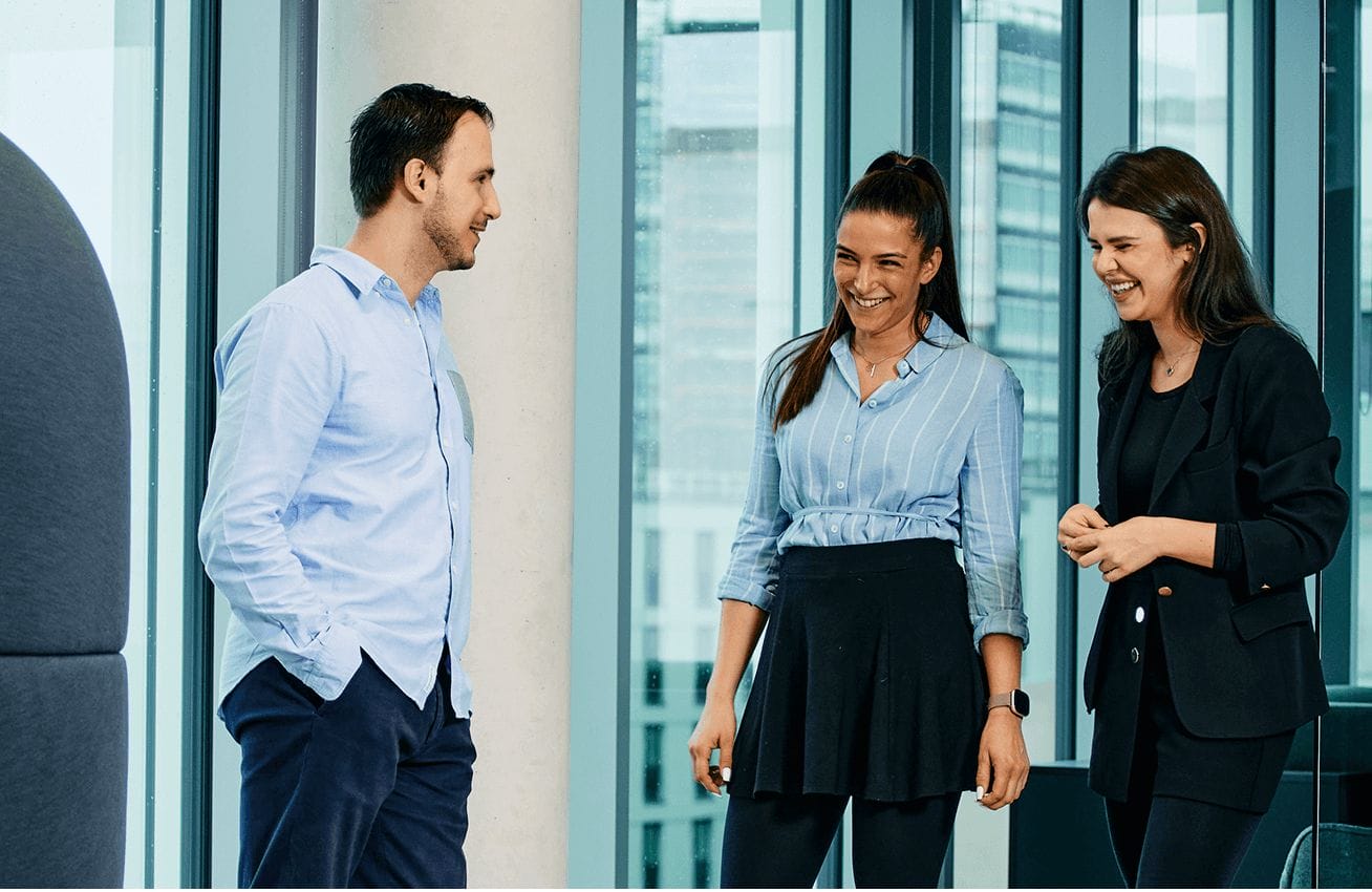 Three people are standing in an office setting, engaging in conversation and laughing. One man is on the left, and two women are on the right. They appear to be enjoying a light-hearted moment, dressed in business casual attire. Large windows are in the background.