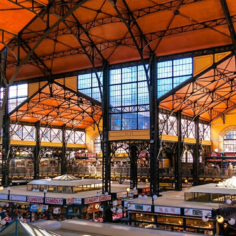 The interior of the central market hall features a high ceiling and multiple stalls selling various goods, all illuminated by natural light through large windows. Expert guides are available to provide insights into local specialties.