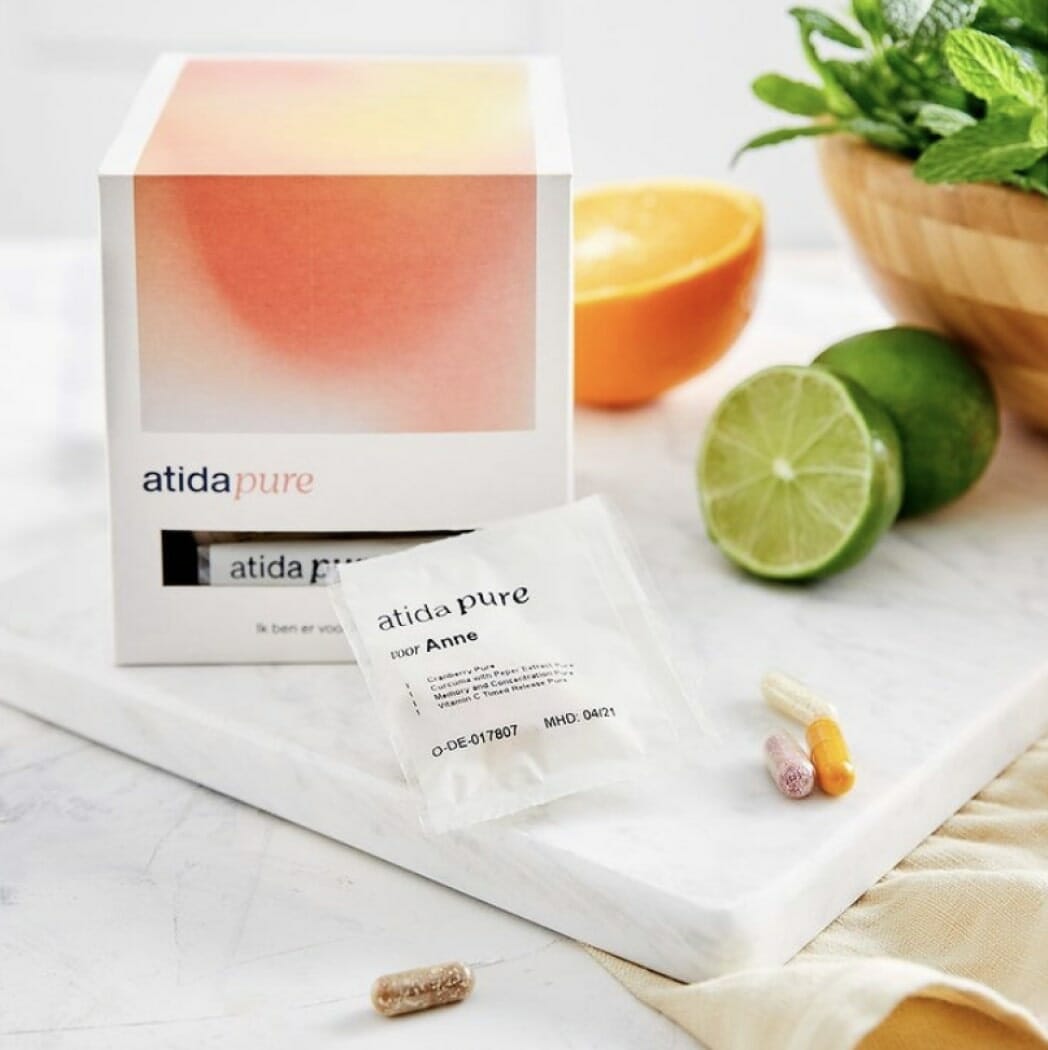 A box of Atida Pure next to an opened packet labeled "Anne" containing capsules and tablets. Nearby are half an orange, a lime, and a bowl of fresh greens on a marble surface, showcasing the natural essence Atida brings to your wellness routine.