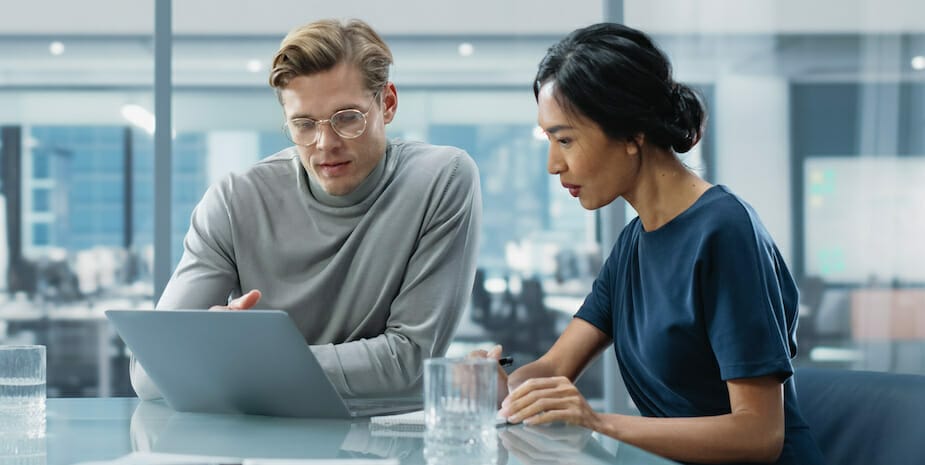 Two people seated at a table in a modern office setting look at a laptop computer screen, discussing the new self-service Request for Quote tool.