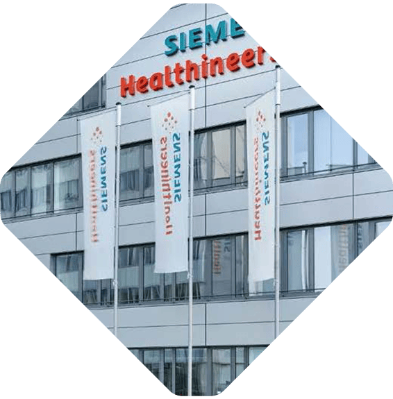 A modern building with the Siemens Healthineers logo displayed at the top. Three flagpoles in front of the building hold white flags, each with the Siemens Healthineers logo. Several windows reflect a cloudy sky. The image is rotated to a diamond orientation.