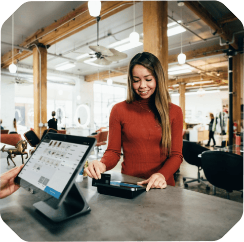 A woman in a red sweater uses a card reader to make a payment at a counter in a well-lit, modern space with wooden beams, hinting at recent expansion. A digital point of sale system is visible.