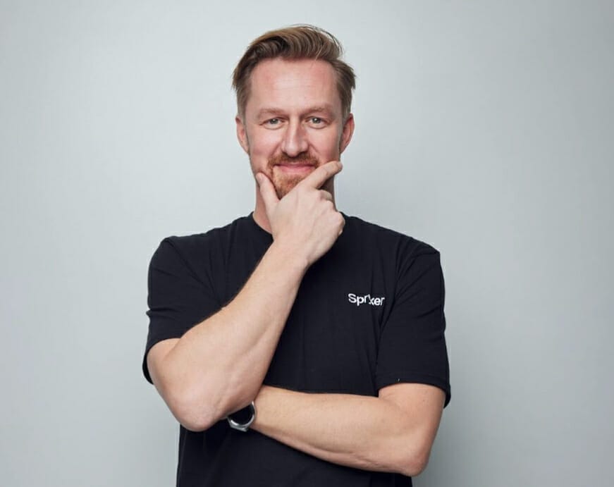 A man with a light beard and mustache, wearing a black t-shirt and a watch, is touching his chin with his left hand against a plain gray background, contemplating the latest trends discussed at eTail Europe.