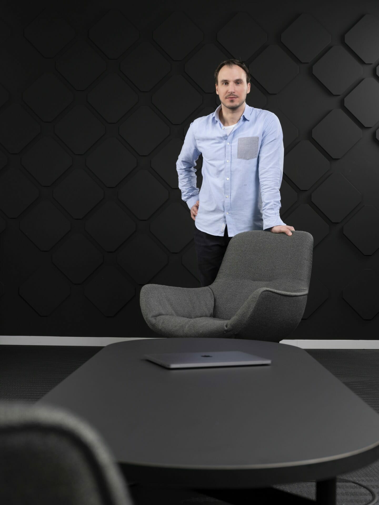 A man stands behind a gray chair in a modern office setting with a black, textured wall in the background. There is a table with a closed laptop, possibly used for developing an app composition platform, in the foreground.