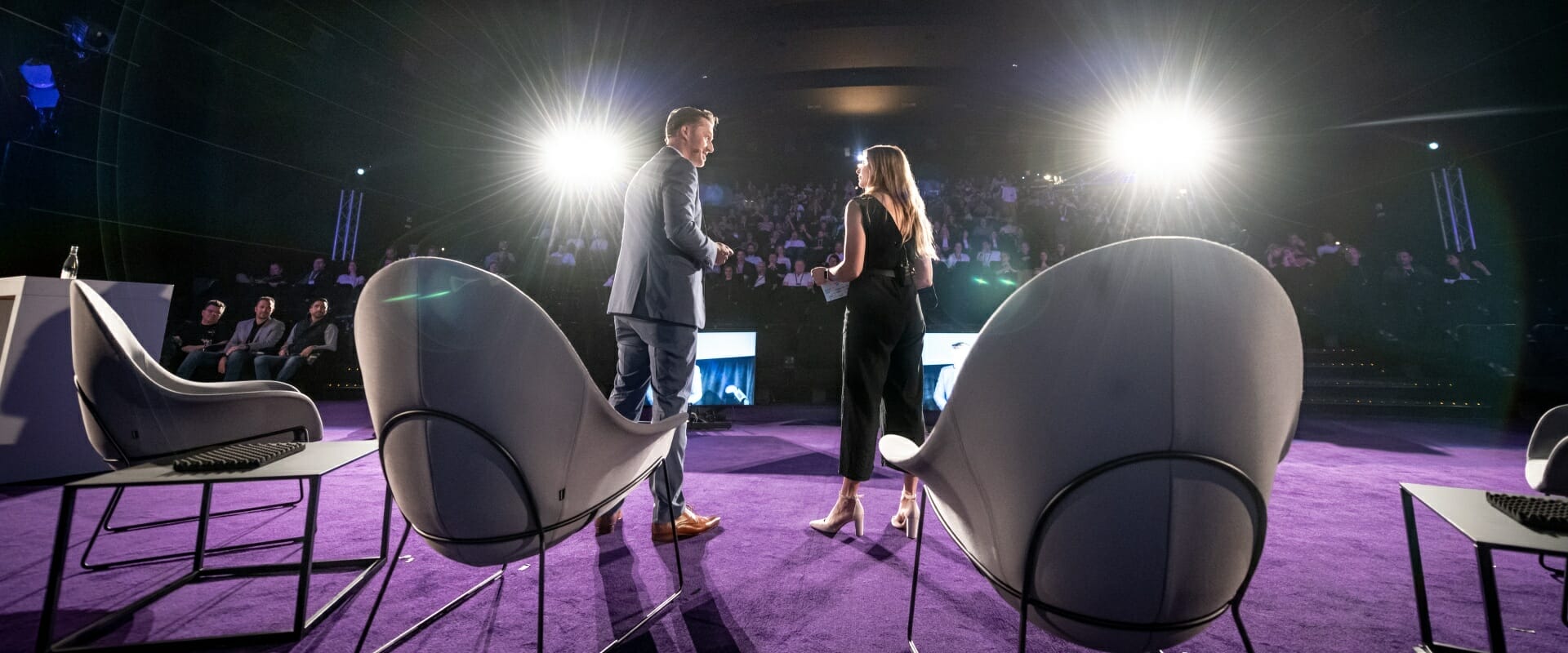 Two people on a purple stage with seated audience, shaking hands in front of two large lights at Spryker EXCITE 2023.